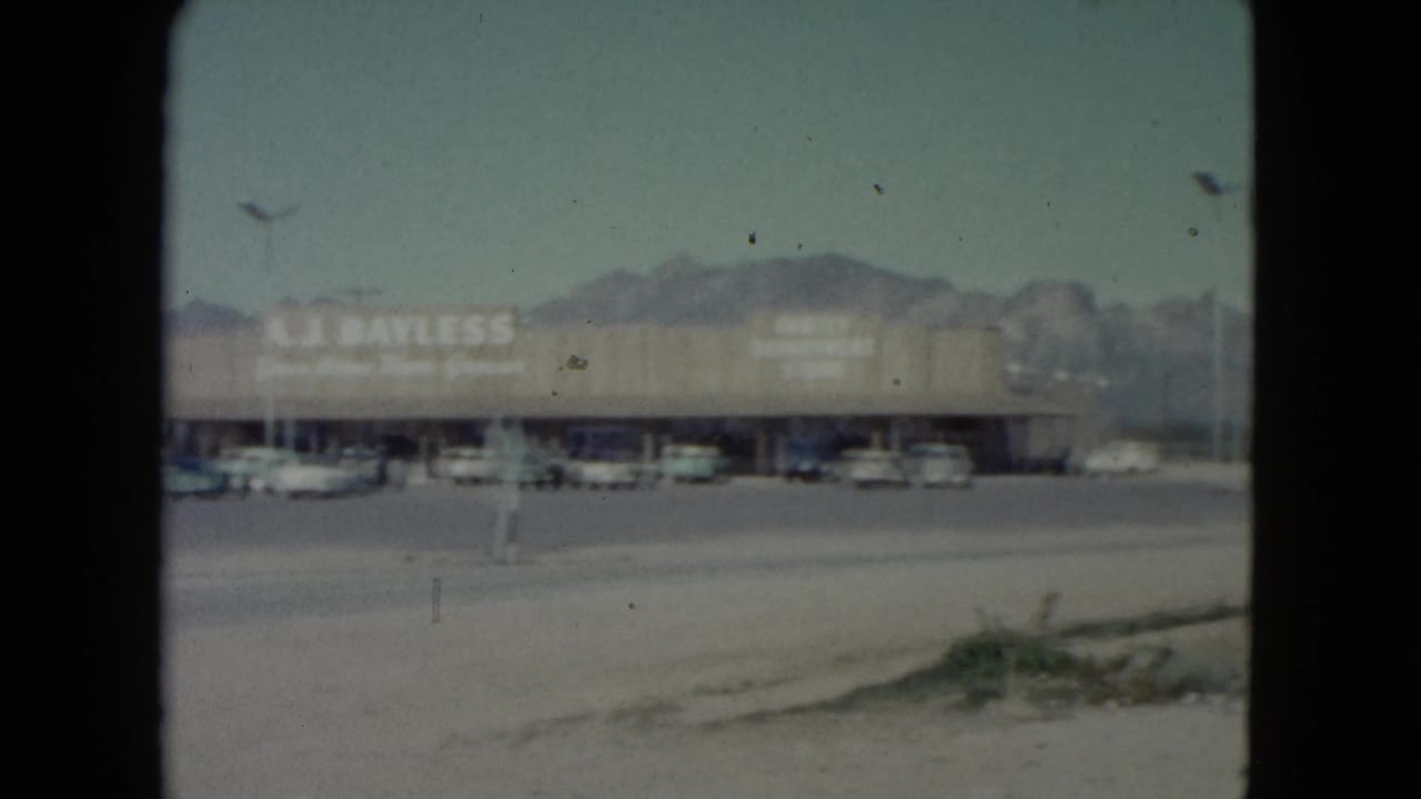 A.J. Bayless storefront across a parking lot with parked cars, light poles, and mountain backdrop.