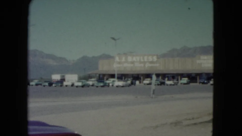 1960 Nevada home-movie view of parked cars outside a long retail building with A.J. Bayless lettering and mountains beyond.