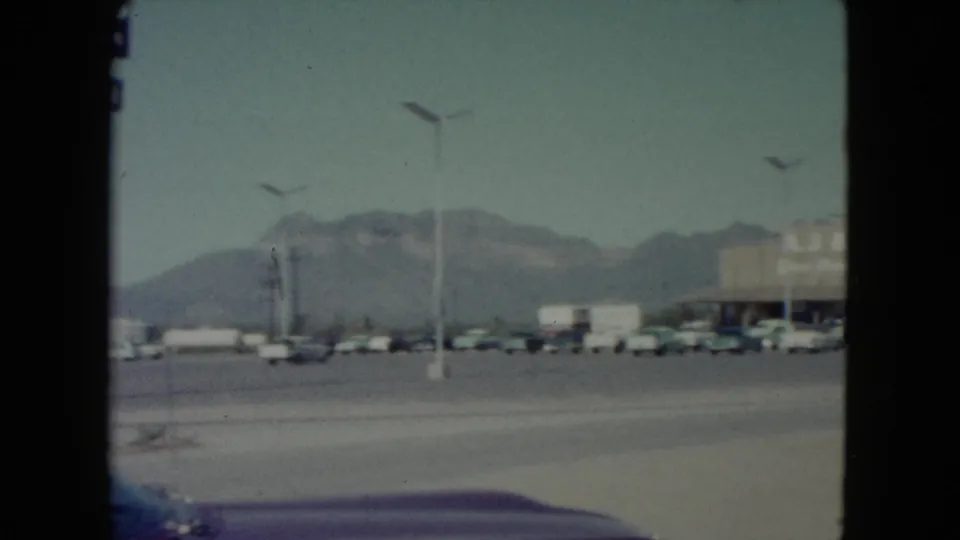 1960 Nevada frame showing the right edge of a retail building, cars in a parking lot, and mountains beyond the road.