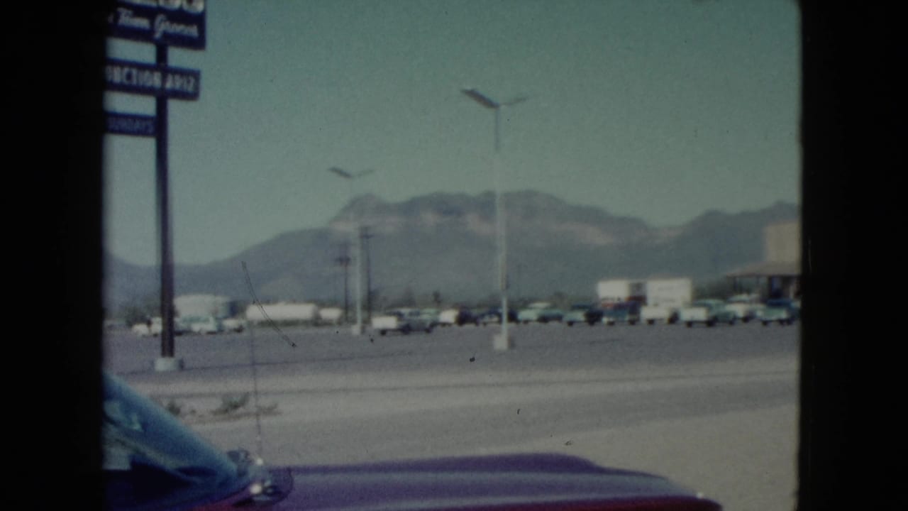 1960 Nevada roadside view from a moving car, showing parked vehicles, a tall commercial sign, hazy mountains, and a broad street.