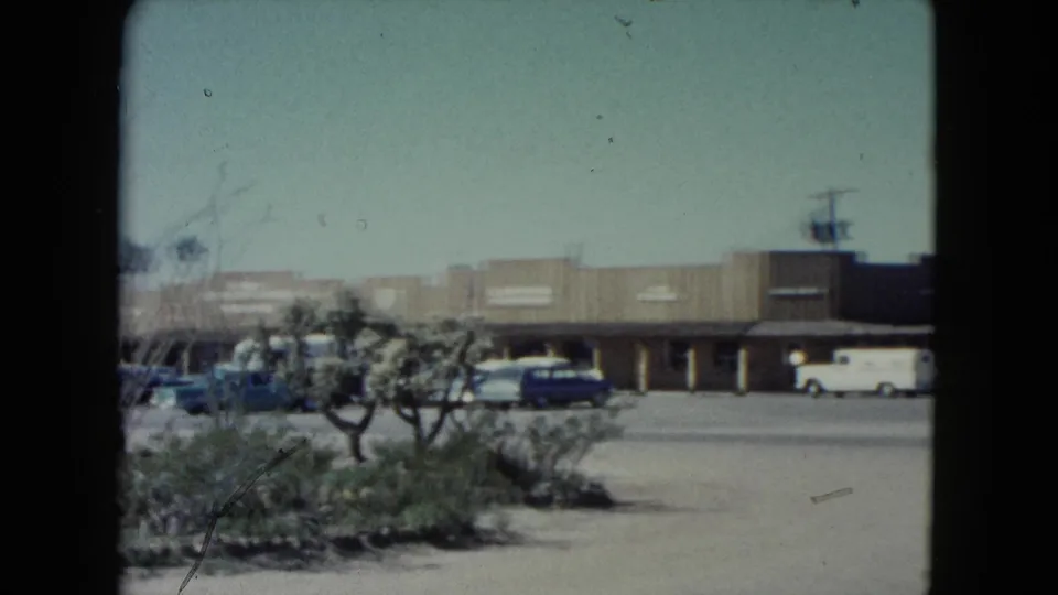 1960 Nevada commercial strip seen from the road, with parked cars, a white vehicle, storefronts, and sparse desert landscaping.