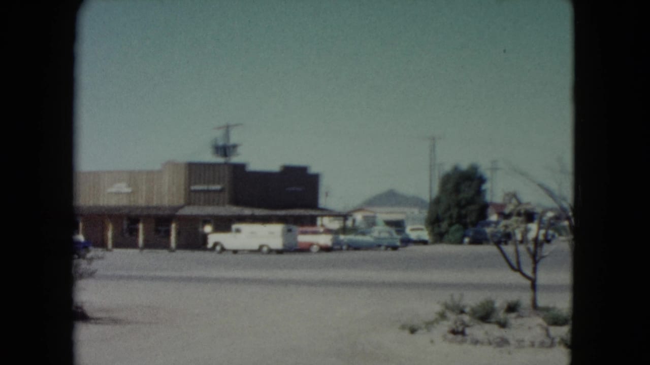 Nevada 1960 home-movie frame showing a low roadside building, parked classic cars, pale sky, and distant mountains.