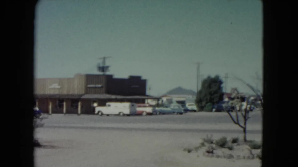 Nevada 1960 home-movie frame showing a low roadside building, parked classic cars, pale sky, and distant mountains.