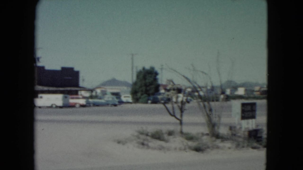 Roadside parking area with low building, cars, small tree, and desert hills beyond.