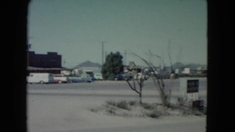 Nevada 1960 frame of a roadside stop with parked cars, a small tree, open pavement, and low mountains in the background.