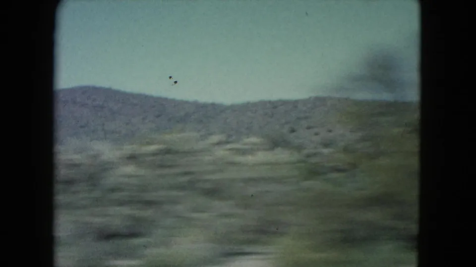 Nevada 1960 moving landscape frame showing blurred desert ground, sparse scrub, and a low mountain ridge under a pale sky.