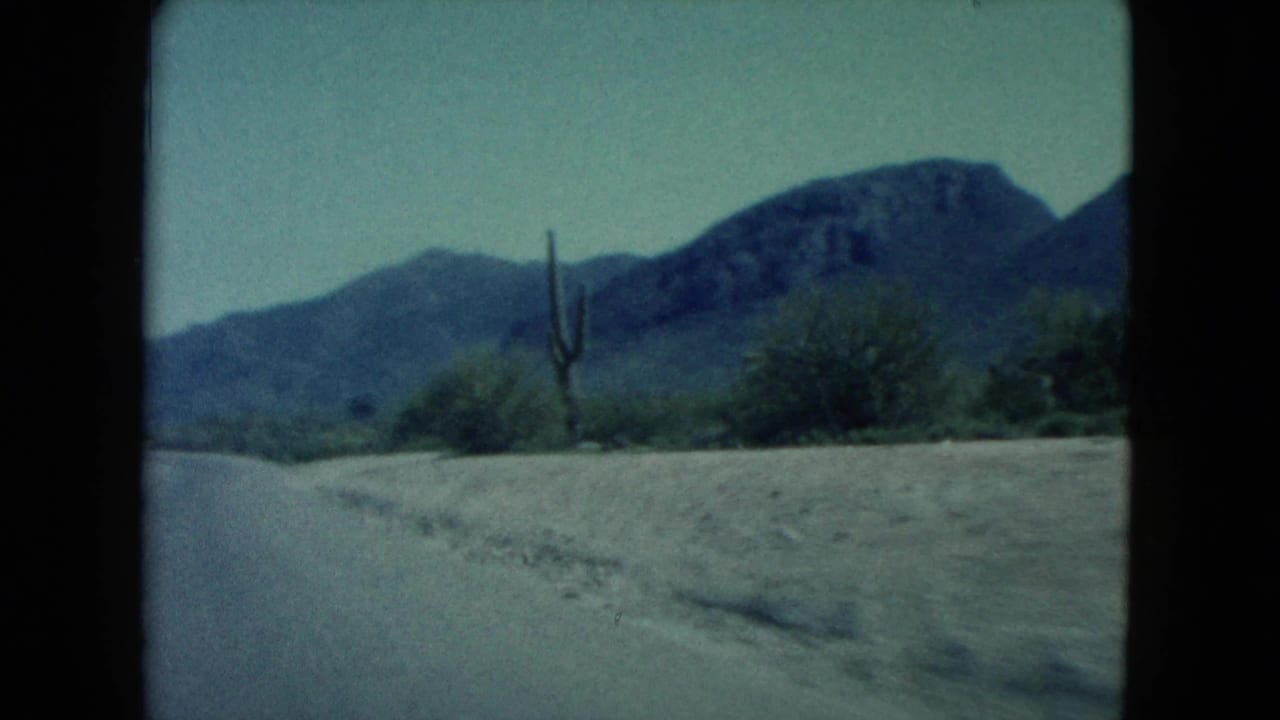 1960 Nevada 8mm frame of a curving road through desert scrub, with tall cactus forms and blue-gray mountains in the distance.