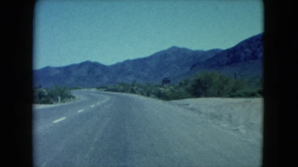 1960 Nevada 8mm frame looking down an open desert road toward low mountains, with sandy shoulders and sparse roadside vegetation.