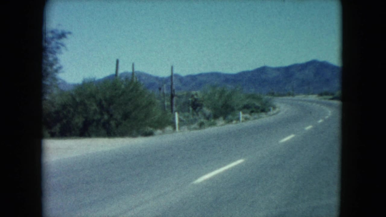 Passenger-view 8mm frame of a Nevada desert road bending past cacti and scrub with mountains in the distance.