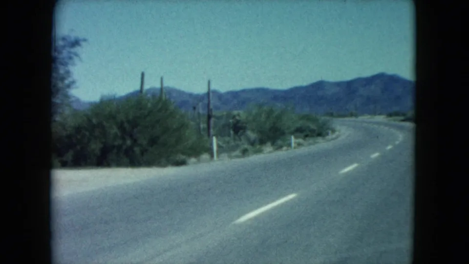 Passenger-view 8mm frame of a Nevada desert road bending past cacti and scrub with mountains in the distance.