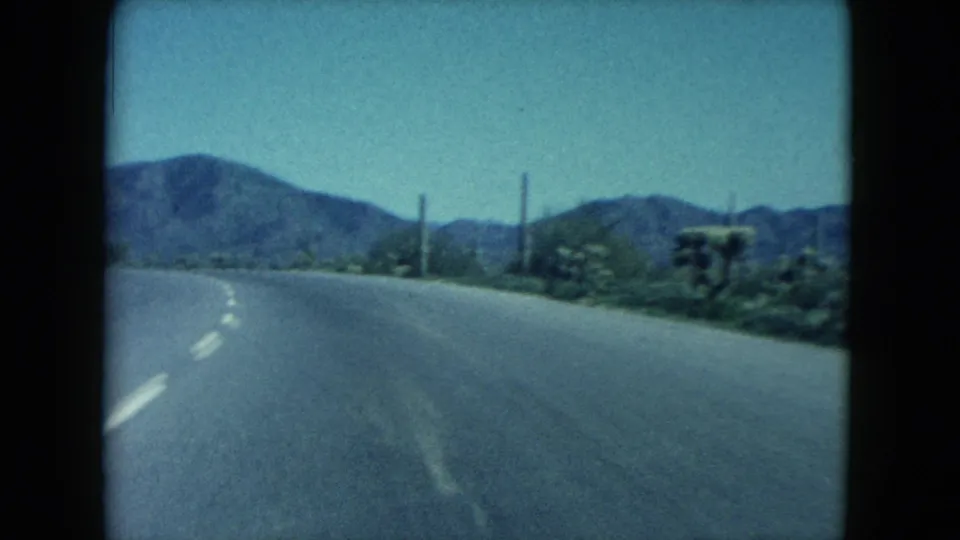 1960 8mm Nevada road-trip view from a moving vehicle, showing a paved highway, roadside brush, utility poles, and distant desert mountains.