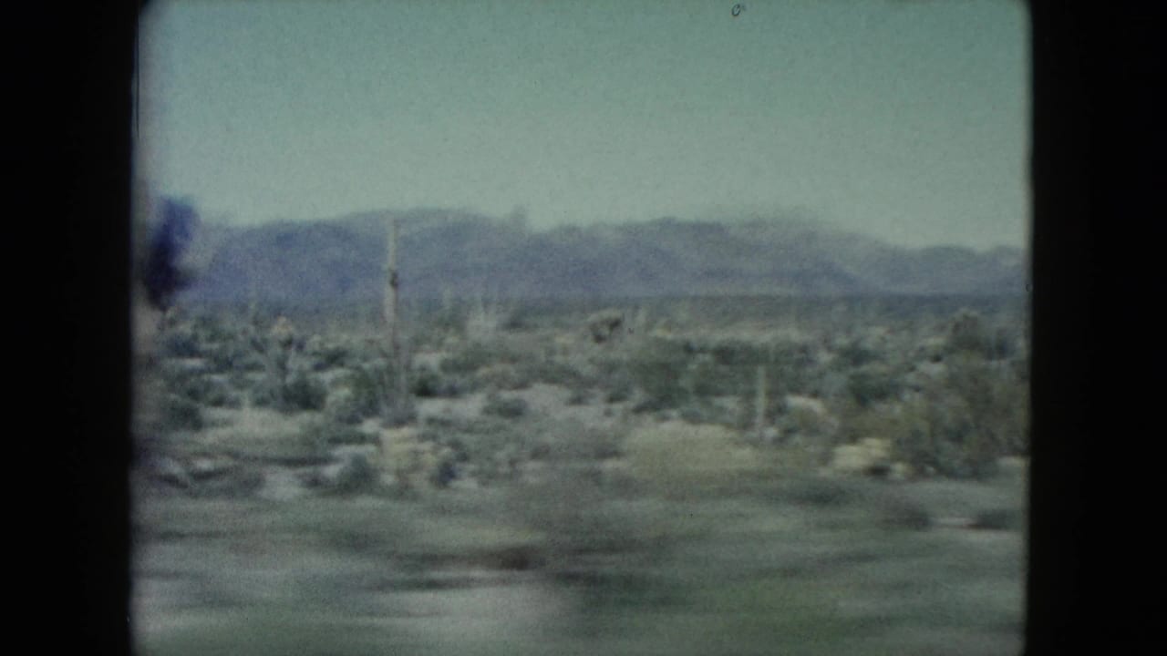 Moving 8mm view across Nevada desert scrub with scattered upright poles, low vegetation, and a hazy mountain range beyond.
