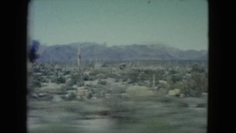 Moving 8mm view across Nevada desert scrub with scattered upright poles, low vegetation, and a hazy mountain range beyond.