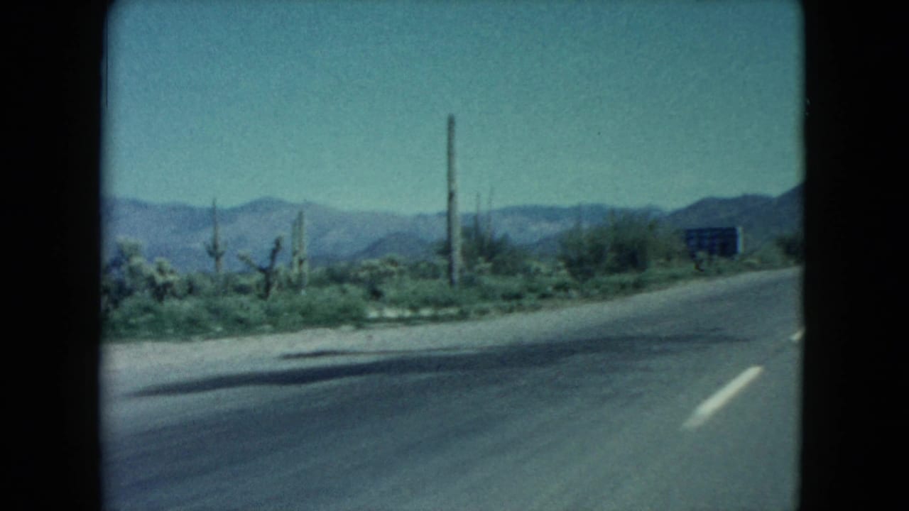 Nevada desert highway scene from 1960 8mm footage, with roadside cacti or Joshua-tree-like plants, a small sign, paved road edge, and distant mountains.