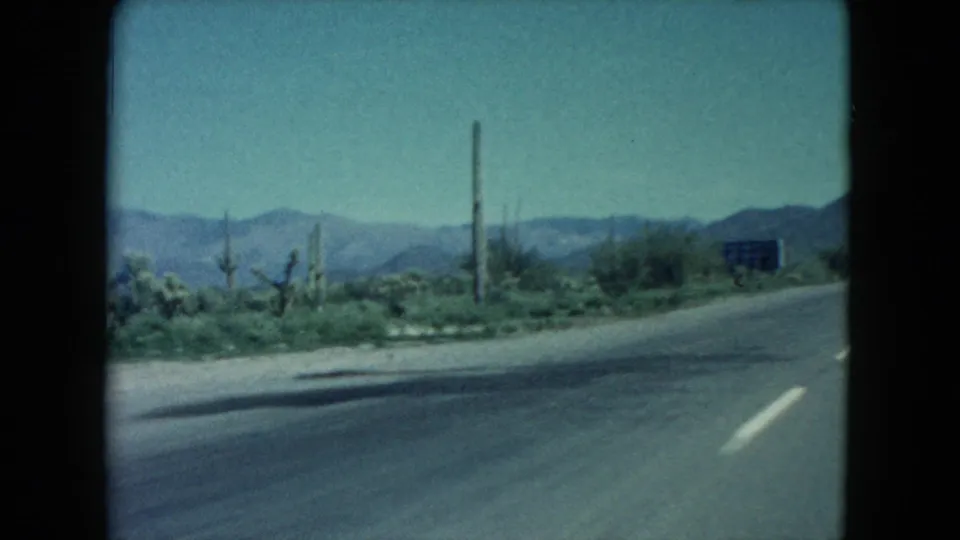Nevada desert highway scene from 1960 8mm footage, with roadside cacti or Joshua-tree-like plants, a small sign, paved road edge, and distant mountains.