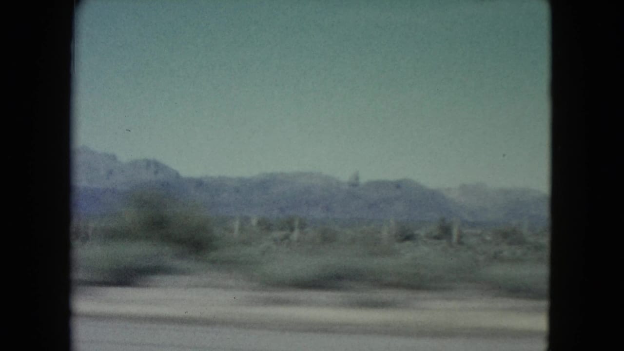 Soft 8mm frame from a moving vehicle showing blurred desert foreground, scattered cacti, low vegetation, and distant Nevada mountains.