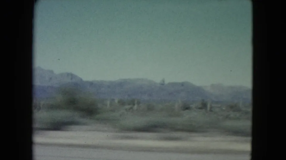 Soft 8mm frame from a moving vehicle showing blurred desert foreground, scattered cacti, low vegetation, and distant Nevada mountains.