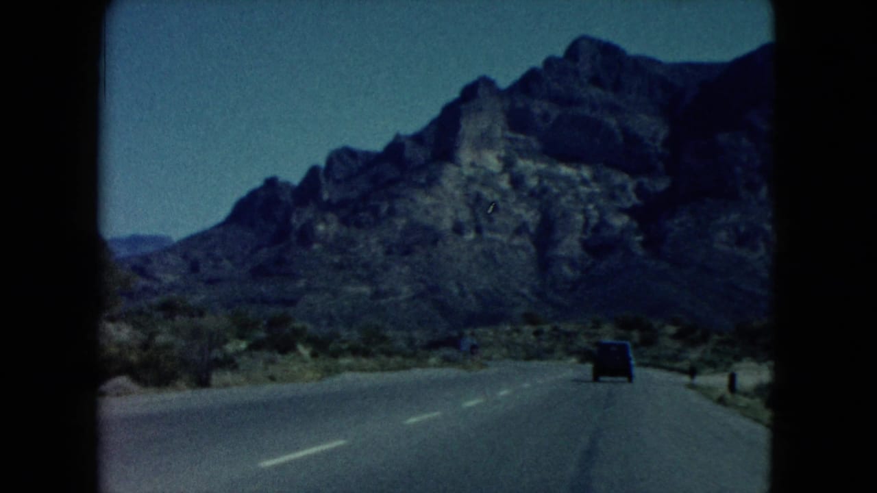 1960 Nevada road-view frame showing a paved highway, roadside markers, desert brush, and a rocky mountain ahead.