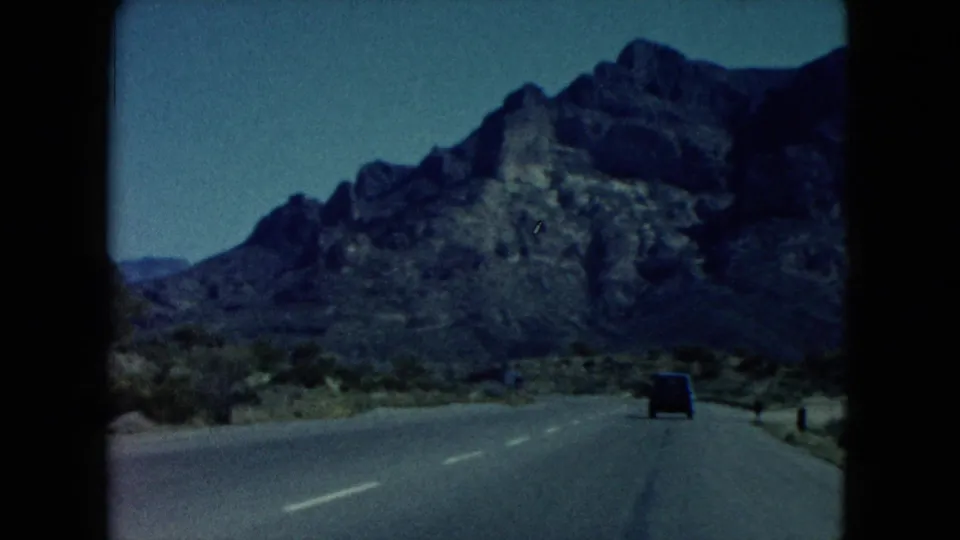 1960 Nevada road-view frame showing a paved highway, roadside markers, desert brush, and a rocky mountain ahead.