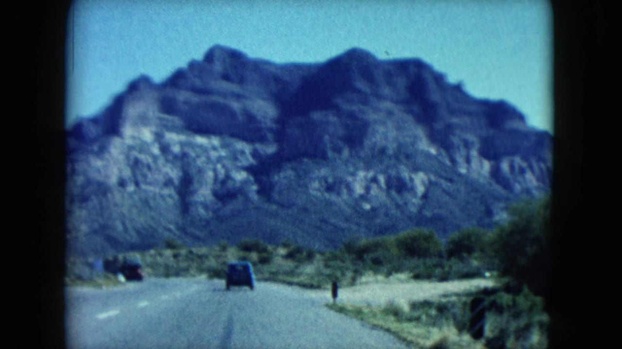 1960 Nevada home-movie view down a desert highway toward a large rocky mountain, with small vehicles visible ahead.