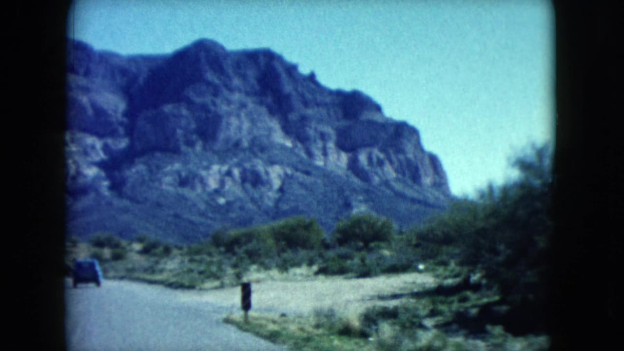 Desert road seen through a car window with mountains and shrubs beyond.