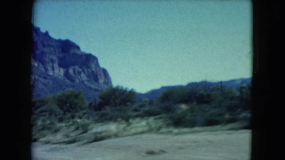 1960 Nevada desert scenery appears through the car interior, with mountains, brush, pale sky, and roadside ground in soft focus.
