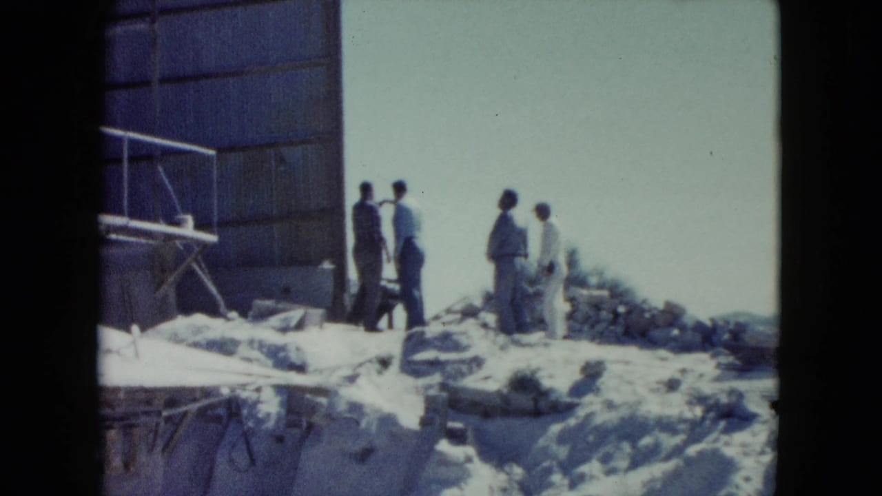 Men in period clothing stand on rocky ground beside a large structure and scaffolding, likely at a Nevada construction site in 1960.