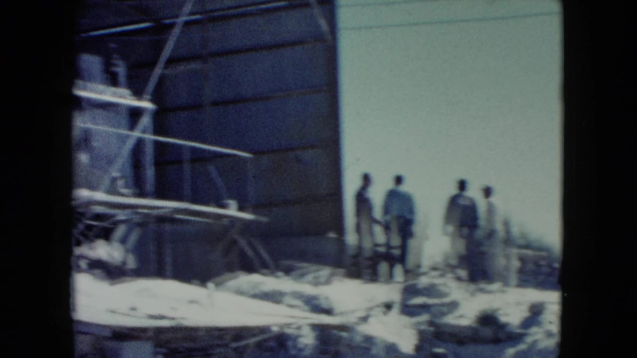 A wider 1960 Nevada worksite view shows men near scaffolding, rocks, and a large industrial-looking structure in bright daylight.