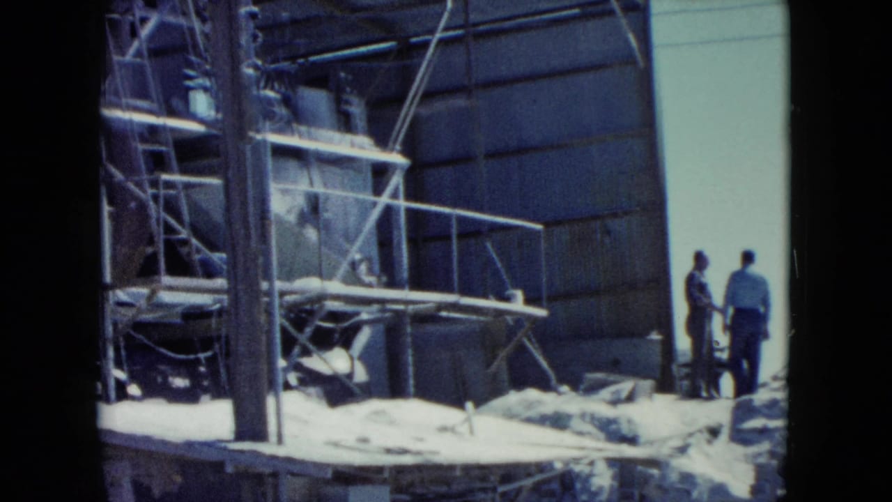 Adult men stand near scaffolding and industrial machinery inside a factory space.