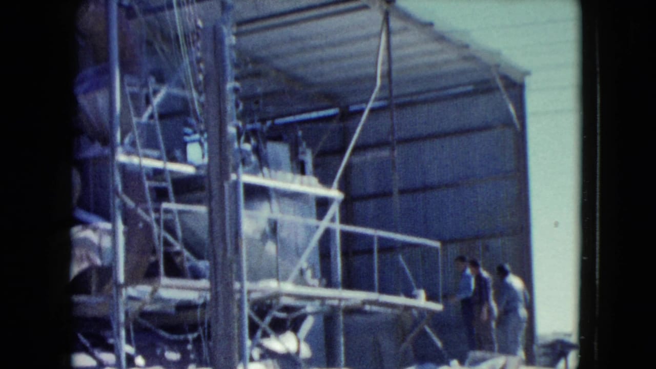 Workers stand near a partially built corrugated metal structure and scaffolding.