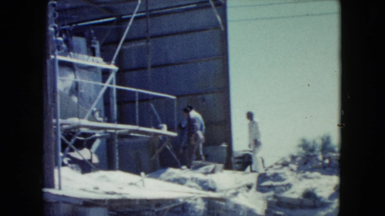 Home-movie frame from Nevada in 1960 showing workers around a corrugated metal structure, scaffolding, debris, and open sky.