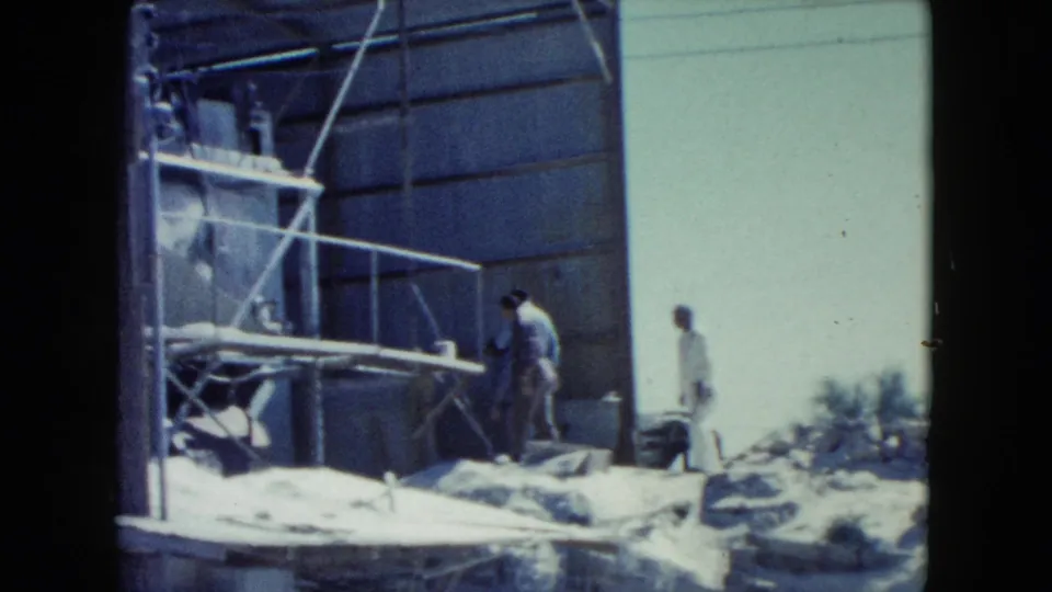 Home-movie frame from Nevada in 1960 showing workers around a corrugated metal structure, scaffolding, debris, and open sky.