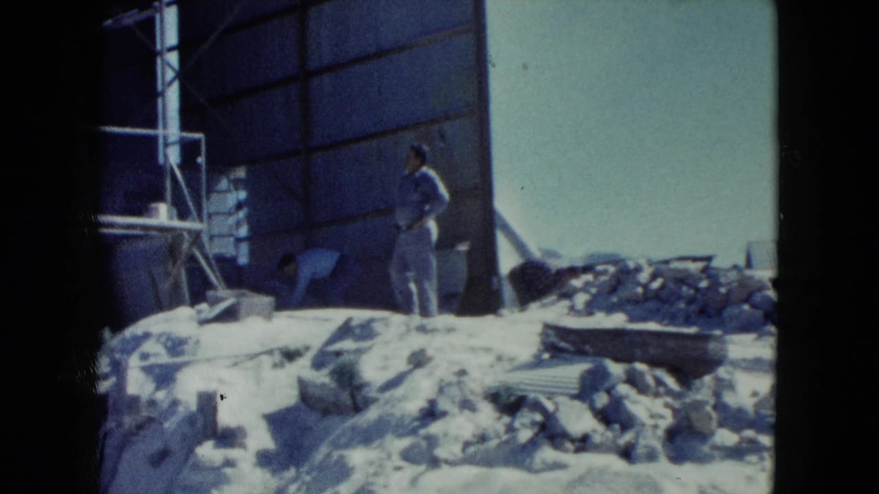 1960 Nevada 8mm frame showing a worker beside a metal building wall, scattered rubble, framing pieces, and bright desert light.