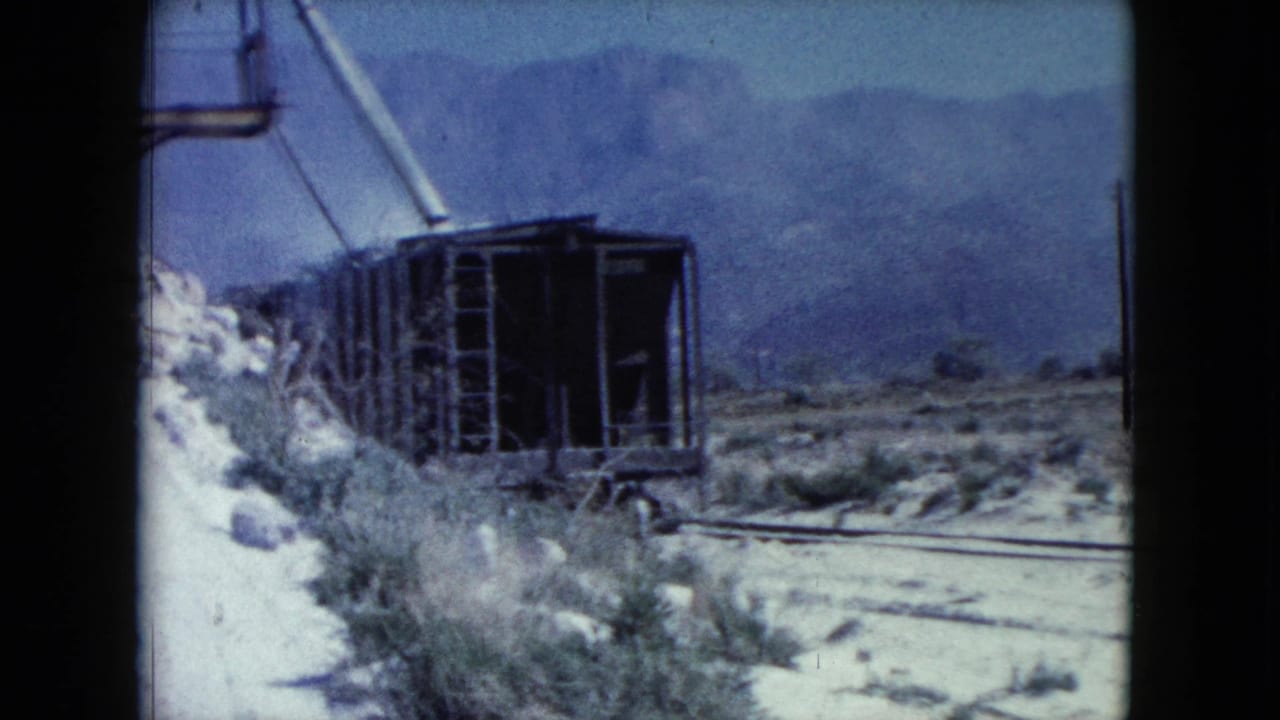 1960 Nevada frame showing a weathered rail car by tracks, rubble, desert vegetation, and hazy mountains beyond.