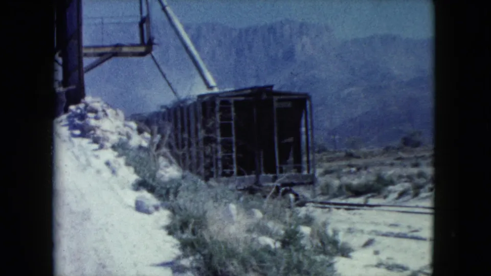1960 Nevada 8mm frame showing an open rail cart near scrub, rock piles, metal equipment, and distant desert mountains.