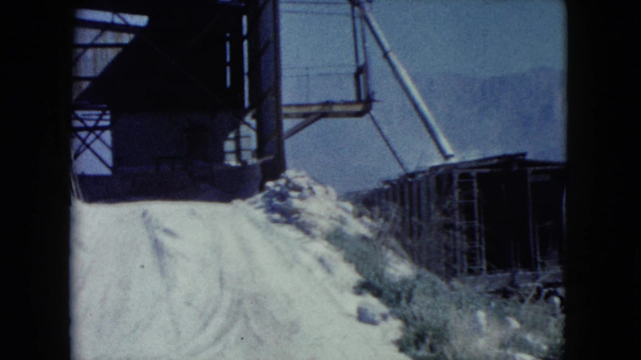 1960 Nevada 8mm view of a pale road, overhead metal structure, open rail cart, scrub, and distant mountains.