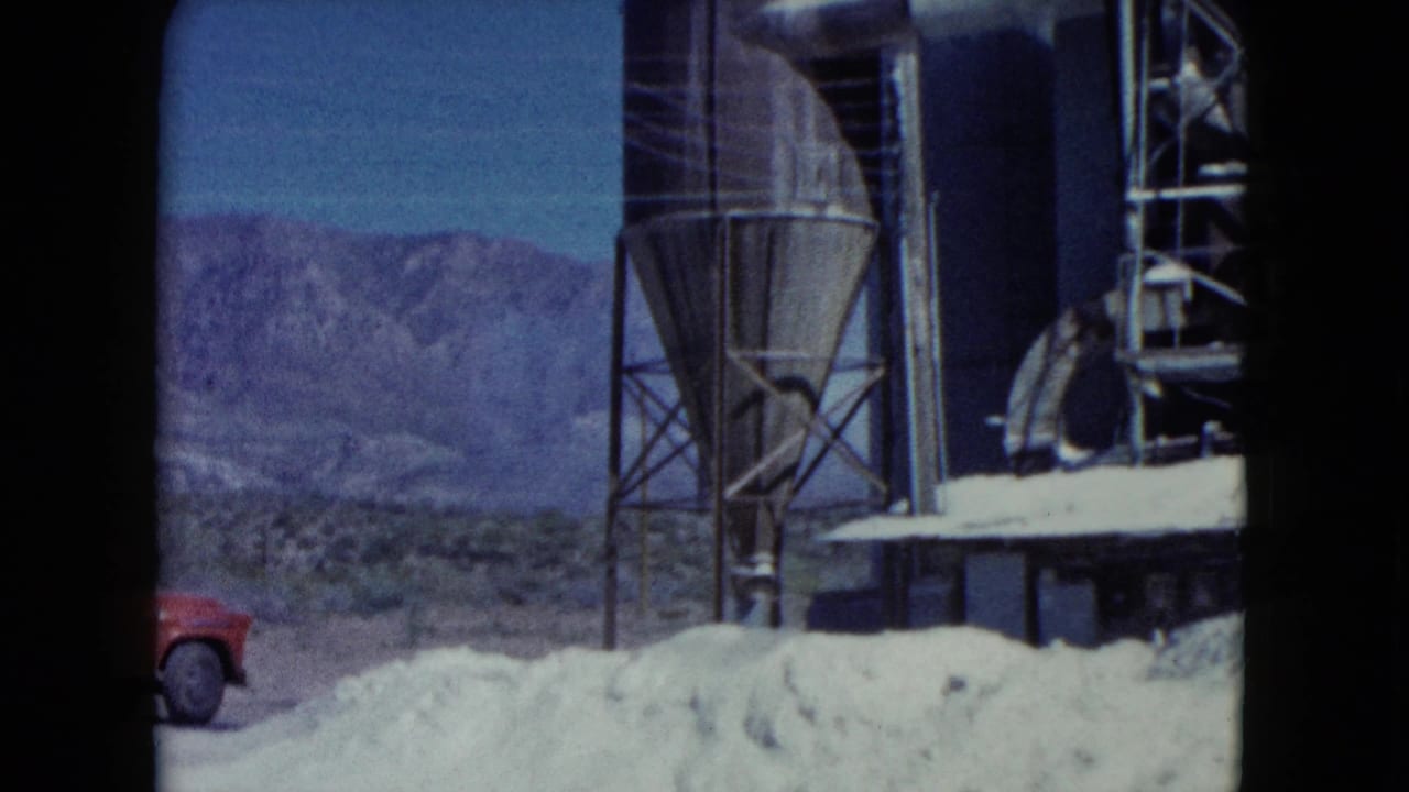 Metal silo and industrial structures beside white material piles in a desert setting.