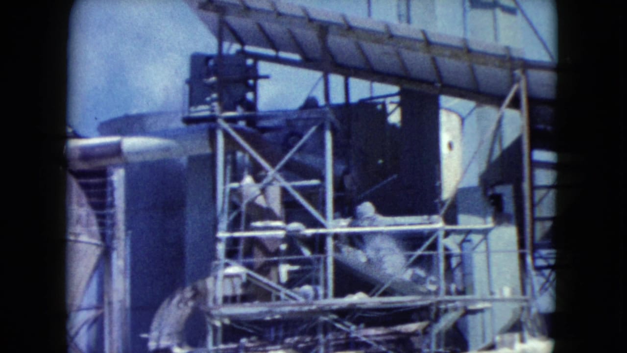 Closer 1960 Nevada 8mm view of conveyor equipment, cross-braced metal framing, pipes, and a light-colored industrial structure.