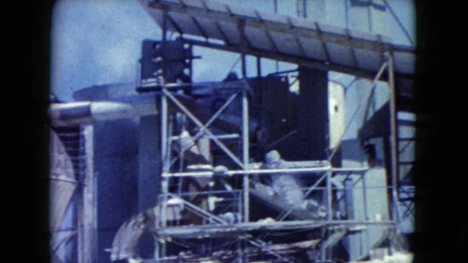 Closer 1960 Nevada 8mm view of conveyor equipment, cross-braced metal framing, pipes, and a light-colored industrial structure.