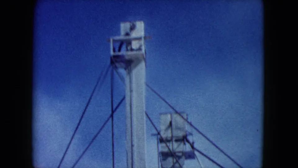 1960 Nevada 8mm frame with a tall white industrial tower, cable bracing, and partial adjacent structure against blue sky.