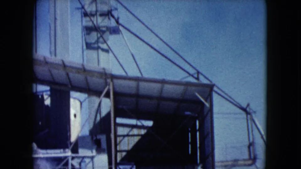 1960 Nevada 8mm frame showing a covered conveyor, white tower edge, steel supports, and open sky at the industrial site.