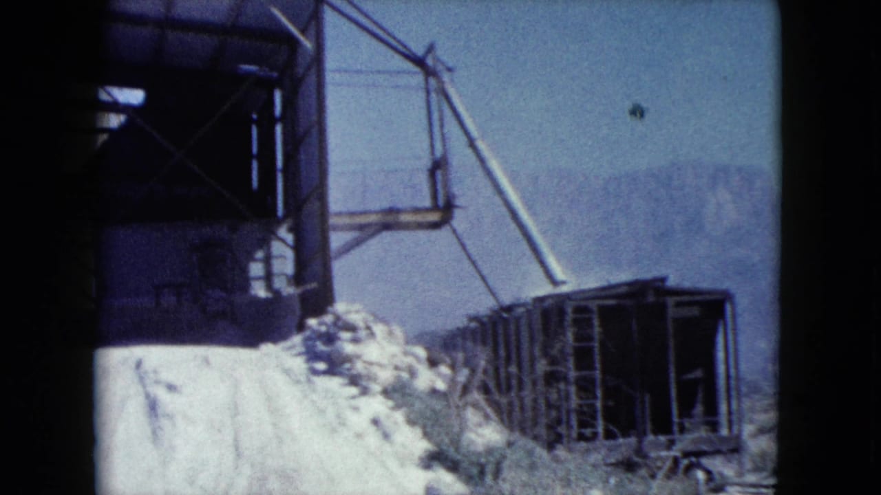 1960 Nevada 8mm frame showing part of the processing structure, a pale material pile, rail cars, and hazy mountains.