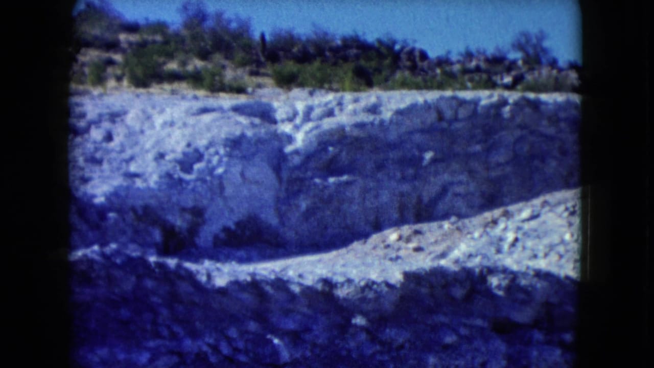 1960 Nevada desert frame showing a pale eroded rock face, scrub vegetation, and a clear blue sky above the cut.