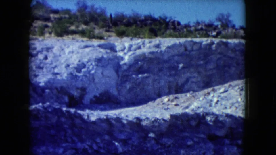 1960 Nevada desert frame showing a pale eroded rock face, scrub vegetation, and a clear blue sky above the cut.