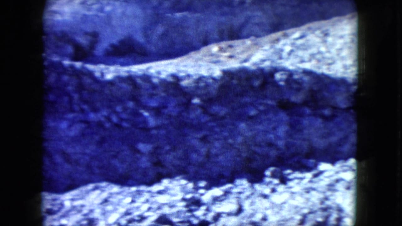 1960 Nevada 8mm frame showing a shadowed eroded bank and bright loose rocks across rough desert ground.