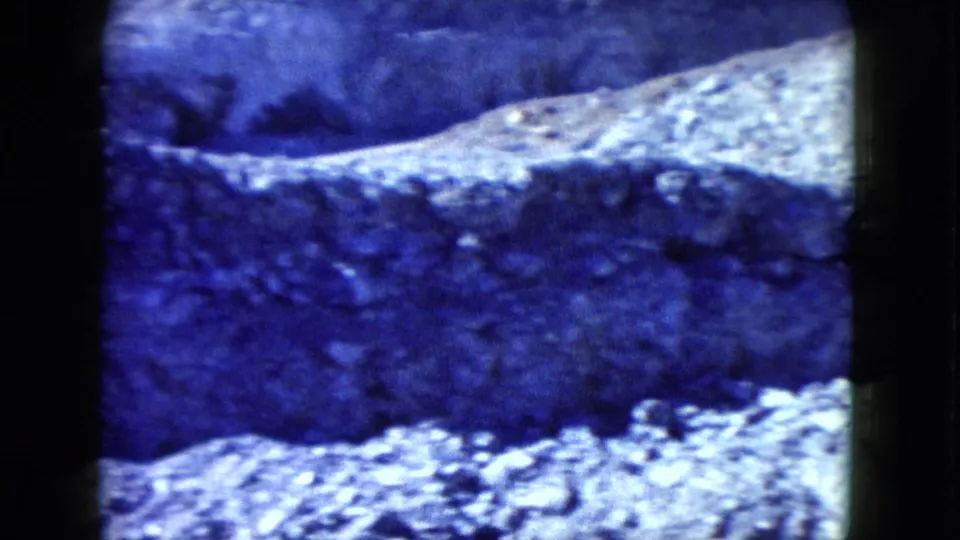 1960 Nevada 8mm frame showing a shadowed eroded bank and bright loose rocks across rough desert ground.