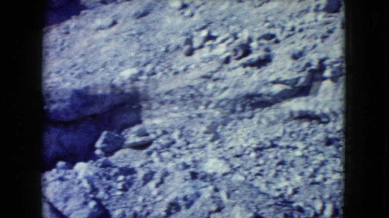 1960 Nevada frame showing a gravelly desert slope, scattered rocks, and eroded pale soil in handheld 8mm color.