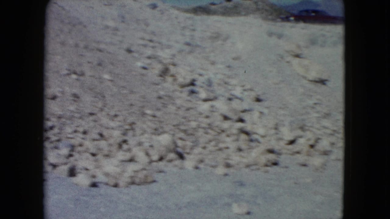 A washed-out 8mm frame shows a dry Nevada desert wash with scattered stones and no visible people.