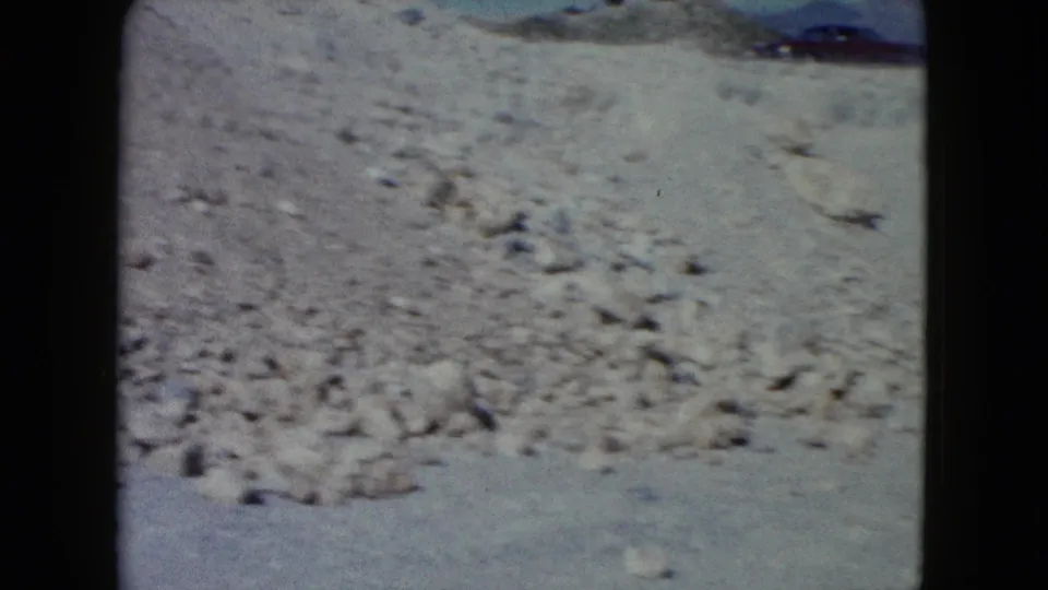 A washed-out 8mm frame shows a dry Nevada desert wash with scattered stones and no visible people.