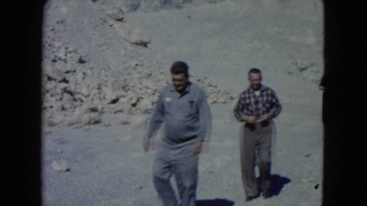 Two adult men, one in a blue shirt and one in plaid, walk through rocky Nevada desert terrain in 1960s 8mm footage.